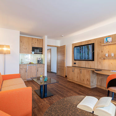 detailaufnahme vom wohnzimmer mit blick über die essecke zur küchenzeile an die tvwand mit hellen holz und orangen möbeln und dunklem vinylboden