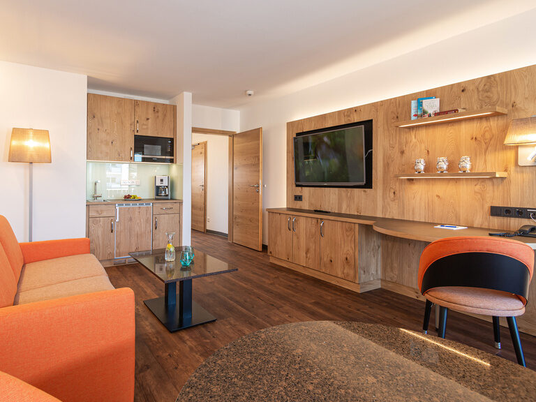 detailaufnahme von der essecke rechte seite tv holz wand aus hellen holz und warmen licht wie die küchenzeile mittig der glastisch und orange schlafcouch