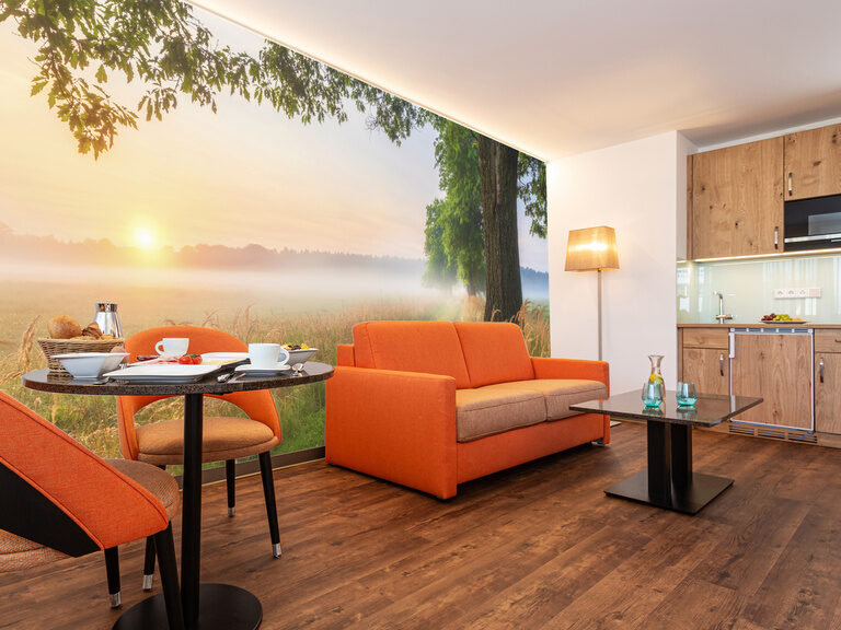 blick ins wohnzimmer auf die fototapete mit sonnenaufgang davor die orange schlafcouch essecke und die küchenzeile mit dunklen vinylboden