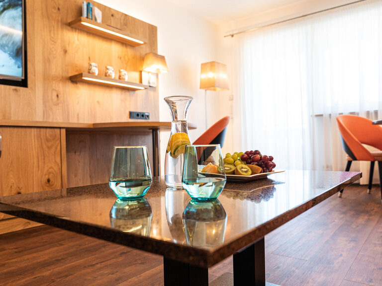 blick durch den raum auf die tvwand mit hellem holz couchtisch mit obstteller recht im bild die essecke mit orangen stühlen