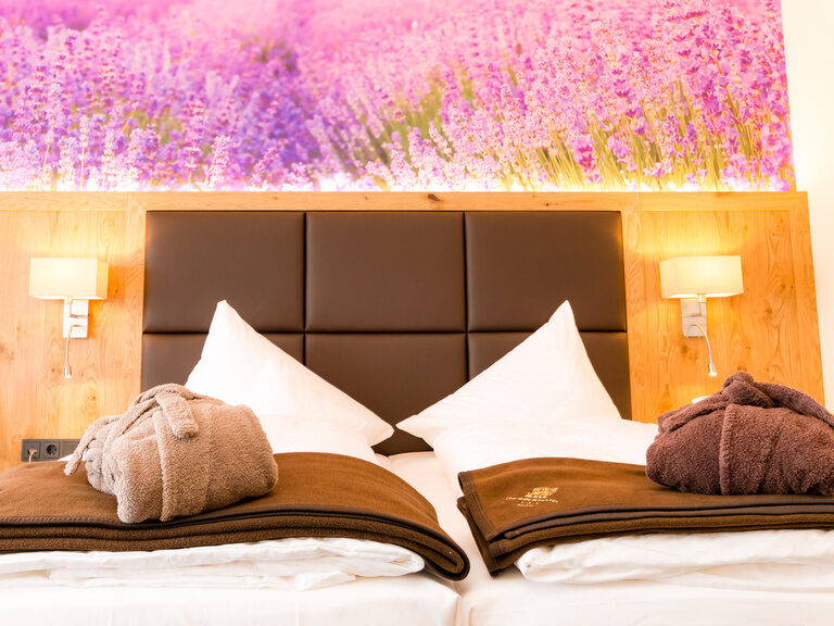 im vordergrund wolldecken in braun auf dem bett bademäntel rückwand in leder links und rechts helle leuchtende lampen darüber eine fototapete mit lavendel