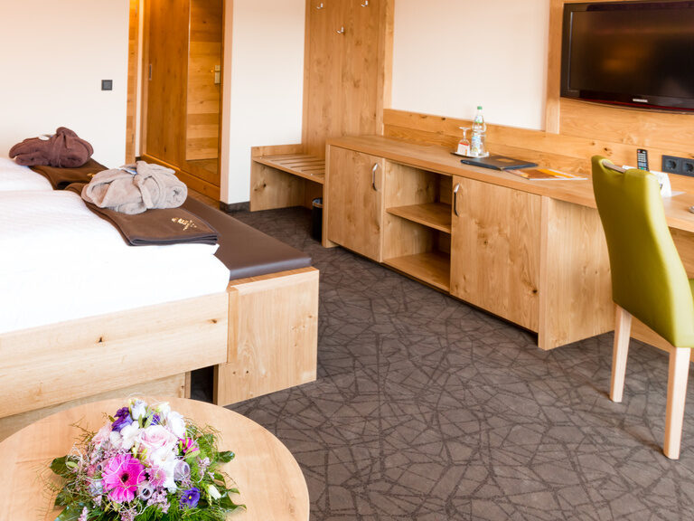 braune wolldecke darüber bademantel auf dem bett davor eine sitzbank mit blick zu tv wand grüner stuhl und helle wohnwand