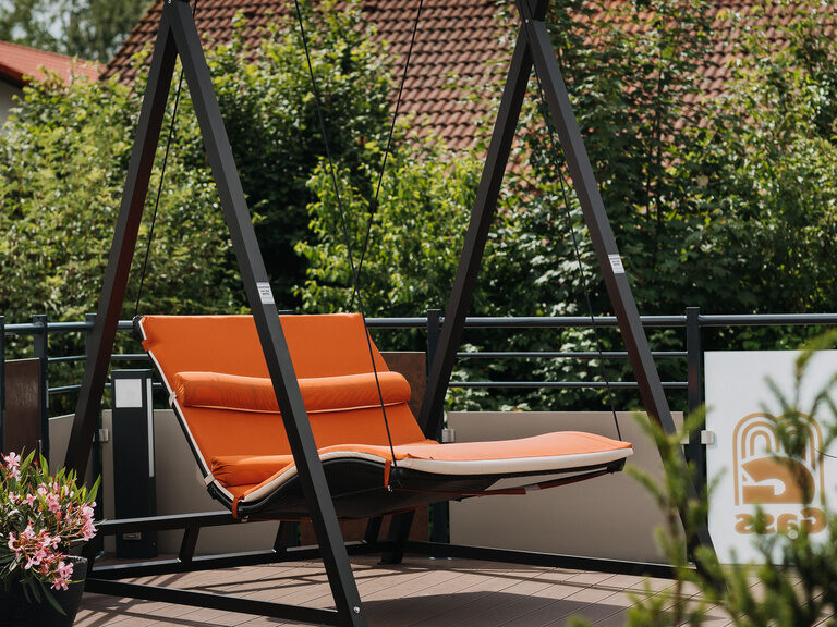 braune schwebeliege mit orangen kissen umgeben von blumen auf unserer aussenterrasse im sonnenschein